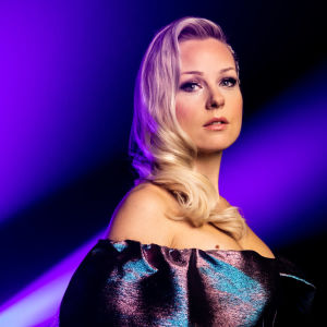 UMK-finalisti Sansa seisoo sinisen taustan edessä ja katsoo suoraan kameraan. Yllään vaaleahiuksisella Sansalla on petrolinvärisenä välkkyvä mekko.