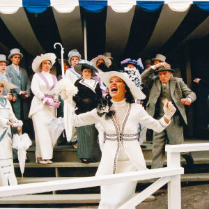 Samppalinnan kesäteatterin My Fair Lady -musikaalin laukkakilpailun kohtaus, jossa naiset hienoissa valkoisissa hatuissaan ja puvuissaan, miehet harmaissa shaketeissaan ja silinterihatuissaan.