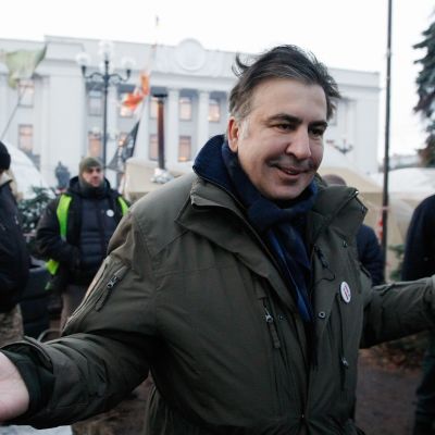 Mihail Saakashvili levittelee käsiään kuvan etualalla, hänen takanaan näkyy muutamia muita miehiä ja Kiovan parlamenttirakennus.