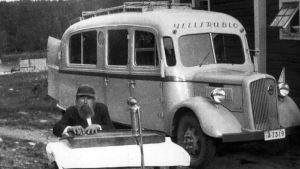 Mies soittaa kannelta ja taustalla "ääniauto Tapernaakkeli, Yleisradion ensimmäinen auto, jossa oli aluksi mekaaniset levytyslaitteet ja myöhemmin magneettiset. Kyljissä kovaääniset radion merkin kohdalla. Katolla kaiteet selostajaa varten".