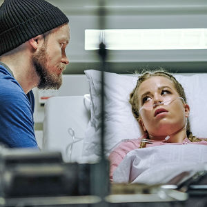 Tyttö makaa sairaalasängyssä happiviiksissä ja katsoo vieressä istuvaa miestä.