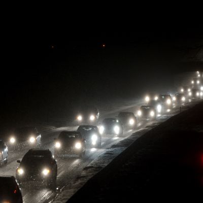 Autoja ajaa pimeässä illassa ja lunta satelee hiljalleen.