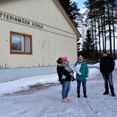 Tre personer står intill ett hus och pratar. Det är vinter. På husväggen står det Petterinmäen koulu.