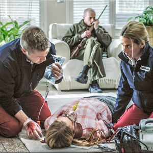 Sairaalasarjan ensihoitajat hoitavat lattialla makaavaa naista. Taustalla sohvalla istuu mies aseen kanssa.