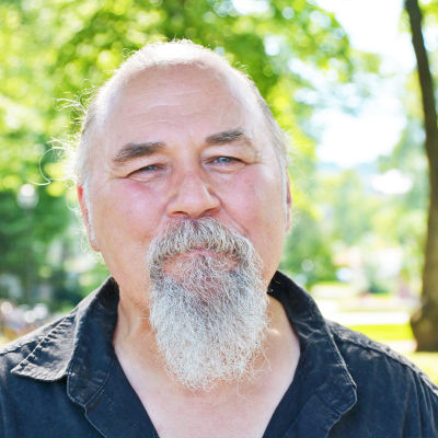 Närbild på en äldre man med grått skägg. Han står ute i en solig och grönskande park och ser glad ut.
