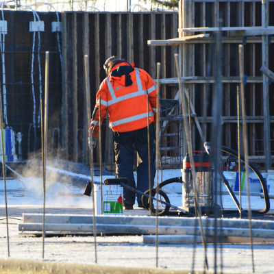 En byggarbetare i röd jacka jobbar på en byggarbetsplats. Han står med ryggen mot kameran och ser ut att hålla i en borste. Bakom honom finns en målfärgsburk och en dammsugare.