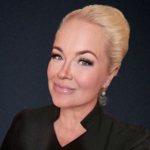 Kati Hoffström anser att man borde testa mycket mera människor än vad man gör i nuläget. en kvinna i blont uppsatt hår och svart skjorta med smink på sig