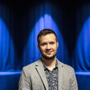 Demian Seesjärvi seisoo teatterin lavalla ja hymyilee kameralle. Taustalla sinisiä teatterivaloja.
