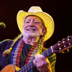 Willie Nelson ler med öppen mun, bär cowboyhatt och spelar akustisk gitarr.