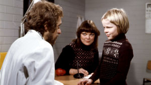 Villahousupakko-elokuvassa 1977 Timo (Kyösti Väntänen) lääkärin (Esa Pakarinen junior) vastaanotolla, taustalla opettaja (Pirkkoliisa Tikka)