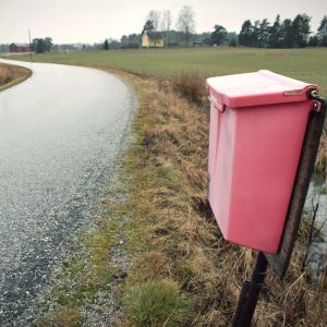 En röd postlåda vid en enslig väg. Ett trähus intill åkerkanten i bakgrunden.