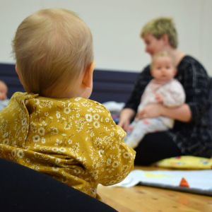 En baby, fotad bakifrån, sitter i famnen på sin mamma och ser mot en annan mamma med baby längre bort.