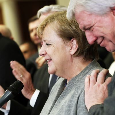 Angela Merkel hymyilee, ympärillä seisovat henkilöt taputtavat.