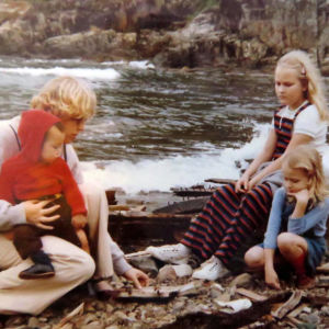 Kivikkoisella rannalla vaalea äiti kiinalaispoika sylissään ja isommat suomalais pikkutytöt istuvat kivellä ja katsovat mitä äidillä on kädessä