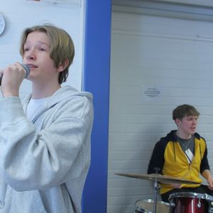 Två ungdomar, pojken längst fram står med mikrofon och sjunger och pojken bakom sitter och spelar trummor.