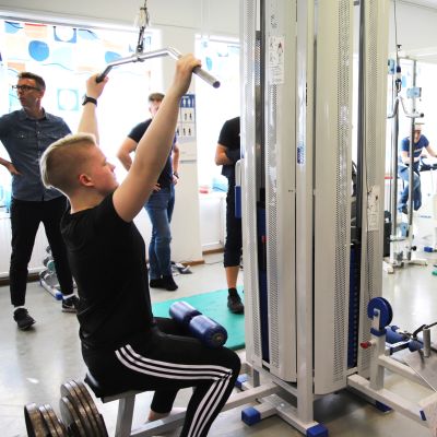 Hepolan koululla personal trainerina työskentelevä Jari Poikela ohjaa nuoria kuntosalilla, kansainväliset vieraat seuraavat kiinnostuneina.