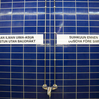 Bild från simhall med texten: Saunaan ilman uima-asua / Gå i bastun utan baddräkt och Suihkuun ennen uintia / Duscha före simningen.