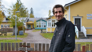 Timo Laine. En man i svart jacka står utanför porten till ett daghem, en träbyggnad i gult och blått. Mannen tittar in i kameran och ler.