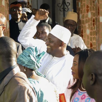 Valkoisiin pukeutunut hymyilevä Adama Barrow heiluttaa kättään. Ympärillä on useita ihmisiä.