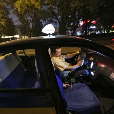 Taksikuljettaja istuu autossaan ja katsoo älypuhelintaan.