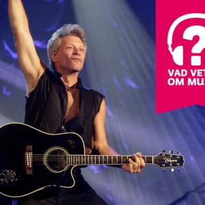 Jon Bon Jovi har en akustisk gitarr kring halsen och han sträcker höger arm rakt upp i luften.