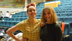 Regissör Lidia Bäck och dirigent Emilie Adolfsson. Två unga kvinnor tittar in i kameran och ler. I bakgrunden synns sittplatser i en konsertsal.