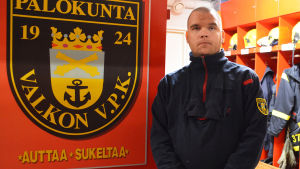 Ray Björklund. En man i blå jacka står i ett omklädningsrum för räddningspersonal. Bakom honom hänger skyddsutrustning på rad och till vänster har han en skylt där det står Valkon VPK palokunta.