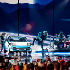 Fyra män klädda i silver står på en scen och framför en sång. På scenen står också en bil.