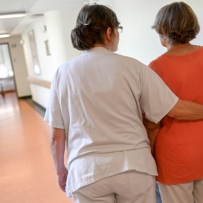 Vanhempi naispuolinen sairaanhoitaja tukee vanhempaa naishenkilöä kävellessä sairaalassa.