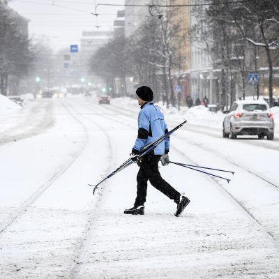 Suksia kantava mies ylittää lumipyryssä katua Helsingissä.