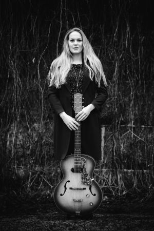Carolina Wendelin fotad av Ian Granström. Kvinna med långt ljust hår ser mot kameran och ler. Hon håller i en gitarr.