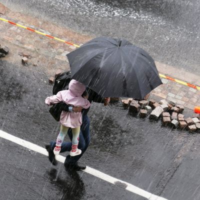 Mies kantaa lasta sateenvarjon alla rankkasateessa Helsingissä.