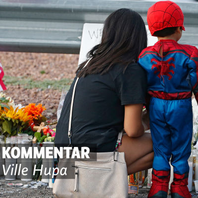 En kvinna och ett barn framför en minnesplats. Det finns mycket ljus och blommor. Text på bilden: Kommentar av Ville Hupa. 
