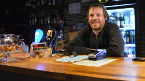 Bybor som inte bor precis invid det planerade smådjurskrematoriet verkar tycka att idén är helt okej. Sami Vuori på byns pub har inte hört några starka åsikter för eller emot. En man står bakom en bardisk. Framför sig har han ett par glasögon och en kassa-apparat.
