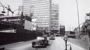 Hötorgsskraporna byggs i centrala Stockholm år 1959. Ett svartvitt fotografi, bilar kör förbi platsen där Hötorgsskraporna i Stockholm håller på att byggas år 1959.