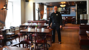 Mika Gehör på Bistro Gustaf. En man klädd i svart står mitt i en tom restaurang. Han ler och håller handen på en stolsrygg.