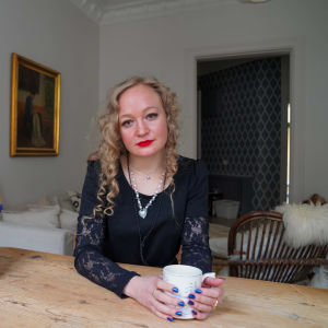 Ida Vainionpää sitter bredvid en bild på sin mamma Riitta Vainionpää vid ett bord.