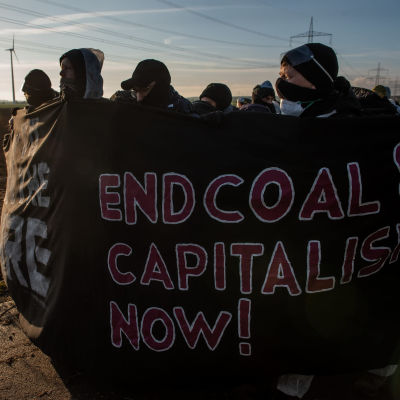 Andreas Malm och en grupp andra människor ute på en klimataktion. Gruppen har masker framför ansiktet och står invirade i en banderoll med budskapet "End coal & capitalism now!"