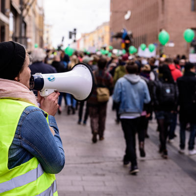 Demonstranter går på Alexandersgatan i Helsingfors. Kvinna talar i en megafon.