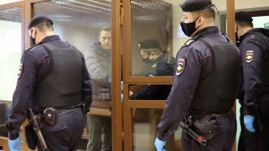 Navalnyj sattes i en låst glasbur i rättssalen på fredagen. Aleksej Navalnyj iklädd en grå jacka förs in i en glasbur av flera polismän.