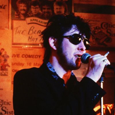 Shane MacGowan har solglasögon, en cigarrett i handen och sjunger i en mikrofon.