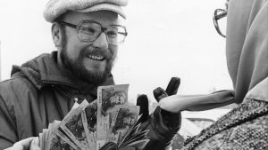 Idel leenden och presenter på jobbet - en kargare och tröttare Karpo på hemmaplan. Hannu Karpo viftar med en bunt sedalr framför en äldre dam på en svartvit arkivbild.