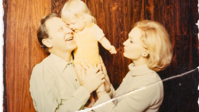 Ett gammalt fotografi på en leende man lyfter ett litet barn medan en kvinna står bredvid och håller händerna om barnet samtidigt.