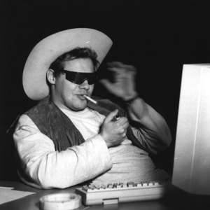 Mies sytyttää savuketta tietokoneen ääressä mustat lasit silmillään.