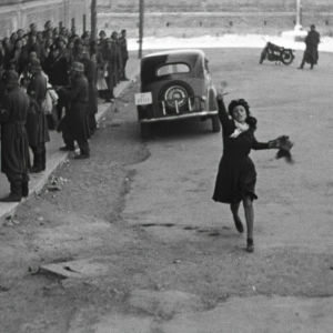 Nainen juoksee kadulla huutaen ja viittilöiden. Kadun laidalla saksalaiset sotilaat pidättelevät väkijoukkoa. Kuva elokuvasta Rooma - avoin kaupunki.