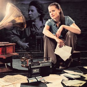 En ung kvinna sitter på en låg pall bredvid en gammal grammofonspelare. I bakgrunden skymtar ett svartvitt foto av Tove Jansson. På golvet ligger gamla papper kringströdda.