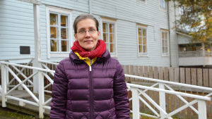 Tuula Heikkilä. En kvinna i lila jacka står utanför ett blått trähus. Kvinnan tittar in i kameran.