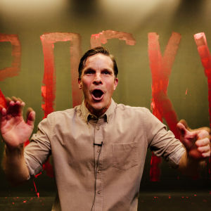 På bilden syns en energisk skådespelare Martin Bahne med armarna i luften och öppen mun. Han är iklädd en beige skjorta och i bakgrunden står det ROCKY skrivet med röda bokstäver.