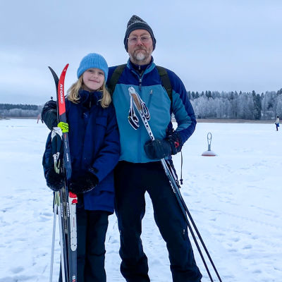 En flicka och en man står på isen med skidstavar i händerna. Flickan håller också i ett par skidor.