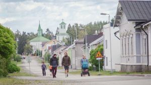 Ihmisiä kävelemässä kadulla, reunoilla puutaloja, taustalla kirkko